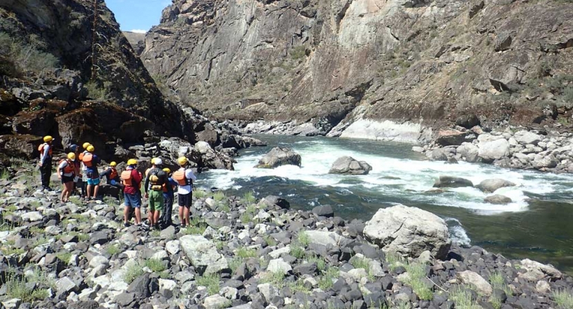 A group of students stand on a rocky shore, observing the river in front of them. The river is framed by high canyon walls. 